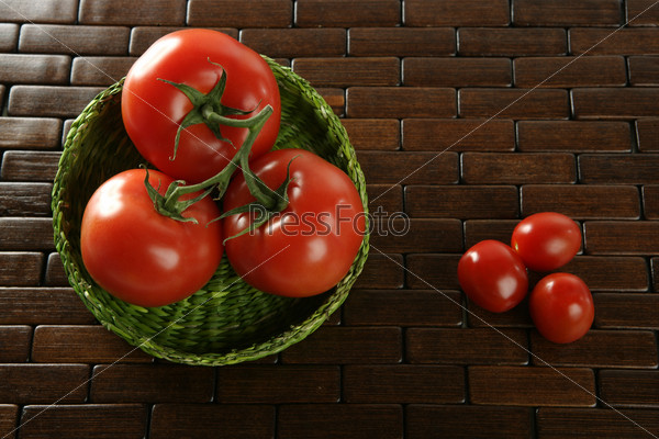 Three little cherry tomatoes and three red big tomatoes in a green basket over wooden table