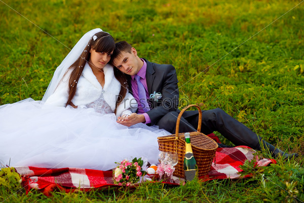 bride and groom wedding in green field sitting on picnic, drink wine from wine glasses at wedding