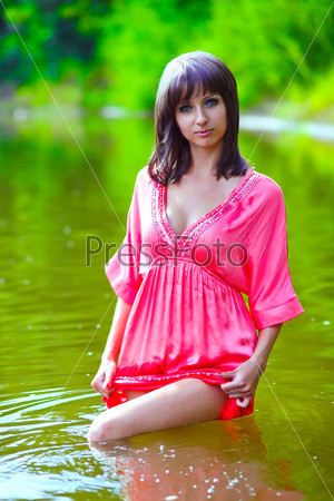 brunette woman model in red dress wet to the waist in the water touches hand of river