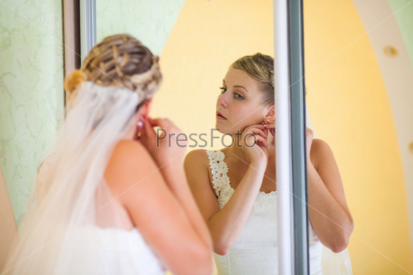 Bride standing in front of a mirror corrects earring wedding\
day