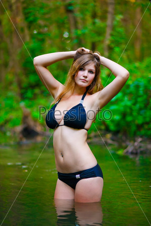 Blonde young woman with big breasts in a black bikini standing on a river in the woods with his hands behind his head