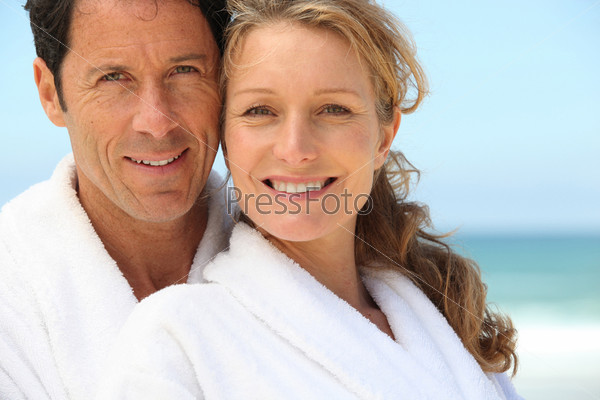 A man and a woman wearing dressing gowns and smiling at us on a beach.