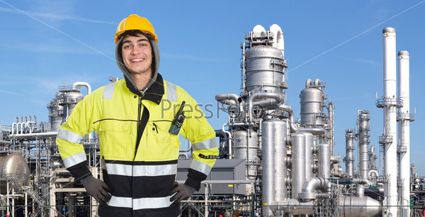 Proud and confident chemical engineer smiling into the camera in front of a petrochemical plabnt, with stainless steel crackers, destillation towers, and a couple of smoke stacks in the background