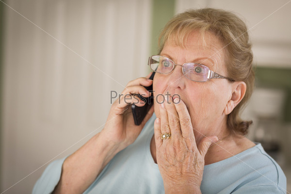 Shocked Senior Adult Woman on Cell Phone with Hand Over Mouth in Kitchen.
