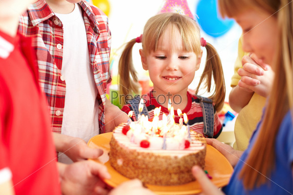Cute girl looking at birthday cake with candles among her friends