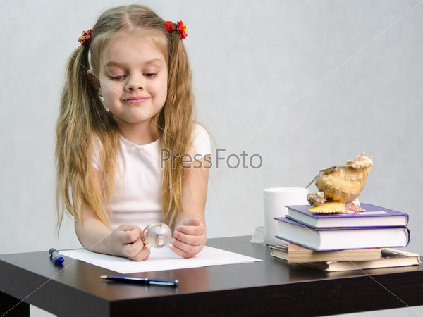 girl thoughtfully and effortlessly turns a small glass globe, sitting at the table. There are tombs of books, sheets of paper, a pen, glass globe, with a glass of tea, sea shell. The image of writer.