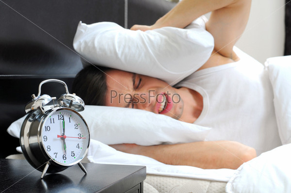 Young male adult stressed by his alarm clock with his head under the pillow in the bedroom