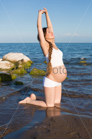 Pregnant woman in sports bra doing exercise in relaxation on yoga pose on sea The concept of health and sport