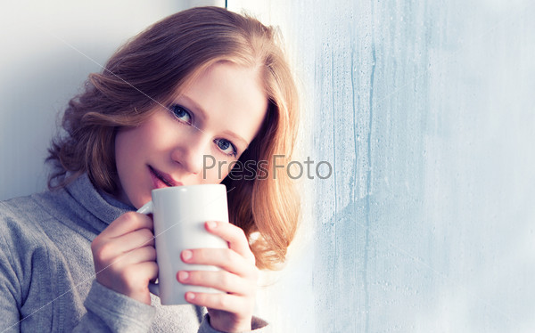 beautiful dreamy young woman with a cup of hot drink coffee at the window in the rain