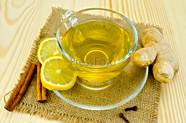 Ginger tea in a glass cup, two slices of lemon, cinnamon, cloves, ginger root on a napkin of burlap and wooden board