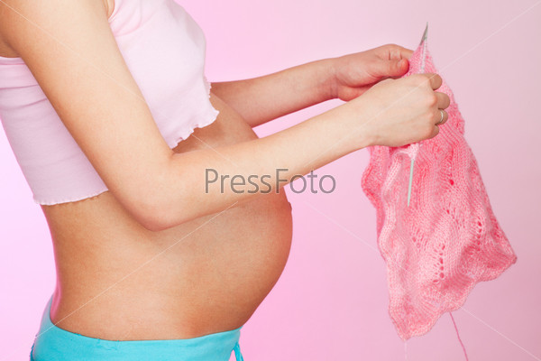 Pregnant woman knitting for a baby on pink background standing in profile, stock photo