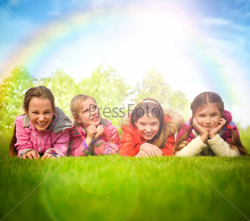 Happy group of girls lying on a green grass. Rainbow and sun overhead