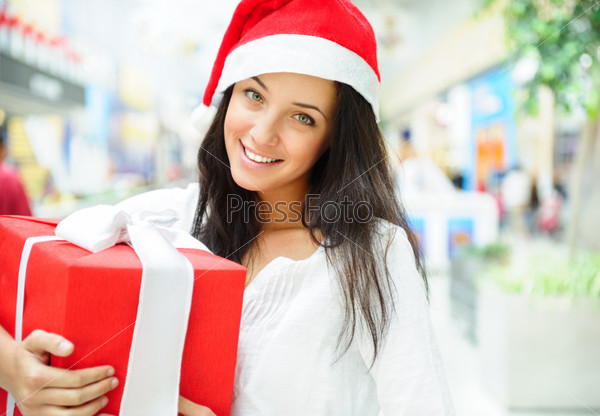 Portrait of young pretty woman wearing Santa Claus helper hat standing inside shopping mall smiling and holding christmas gift