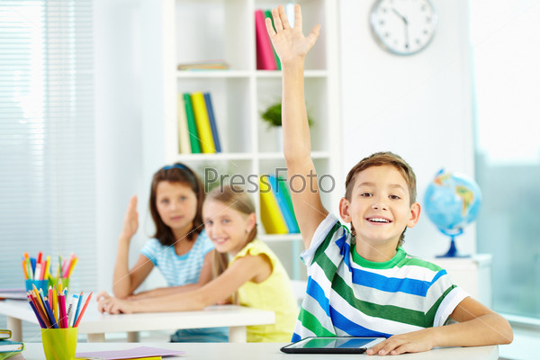 Portrait of clever schoolboy raising hand at workplace with two classmates behind
