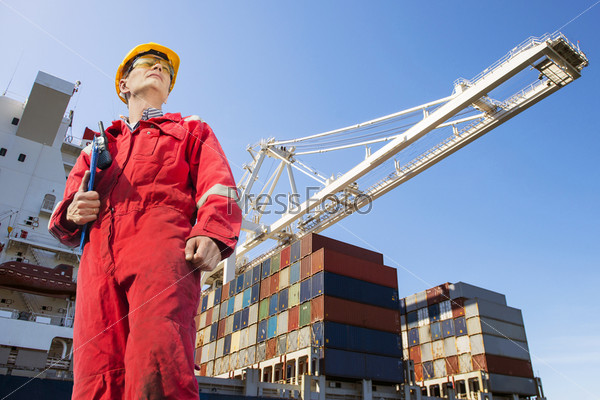 Harbor master with clipboard, overalls, hard hat and safety glasses standing in front of a large container ship being unloaded