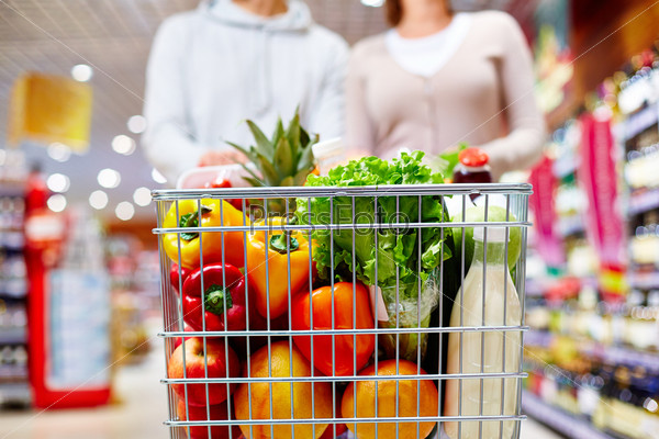 Image of cart full of products in supermarket being pushed by couple
