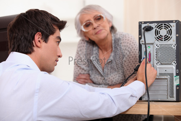young man wearing a shirt is plugging cables on the back of a computer, an old woman is watching him