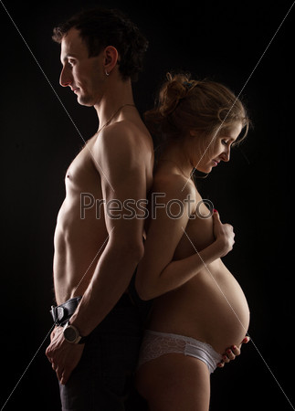 Portrait of young pregnant couple standing back to back against black background