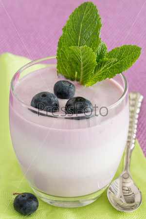 Glass of fruit yogurt dessert with blueberries, spoon and mint decoration, on purple and green background