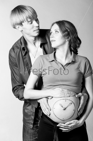 clock. Man is embracing stomach of a pregnant woman on gray background with painted watch