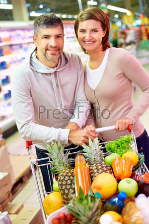 Image of happy couple with cart full of products looking at camera in supermarket