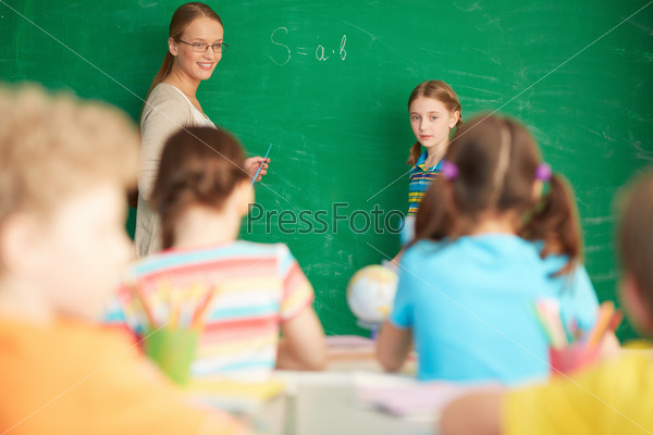 Portrait of smart teacher and schoolgirl standing by blackboard and looking at schoolkids in classroom