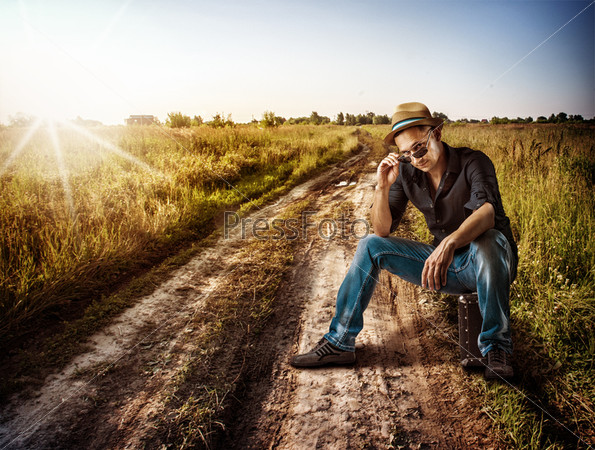 Young handsome man traveler sitting on his suitcase on a dirt road outdoor