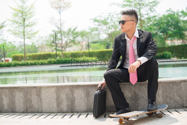 Young businessman in suit sitting outdoors with a briefcase and a skateboard
