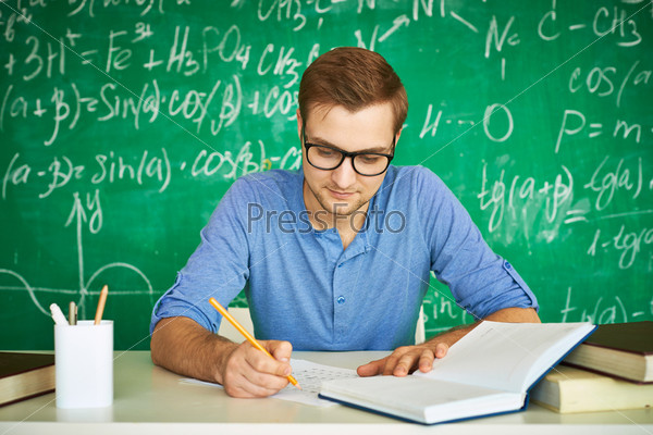 Portrait of handsome student carrying out graduation test on background of chalkboard, stock photo
