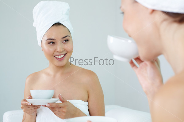 Girl in bath towel talking to her friend in spa salon while drinking tea