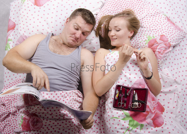 Husband and wife are lying in bed. Wife handles nails manicure nail file, husband reading a magazine and shows an interesting picture wife