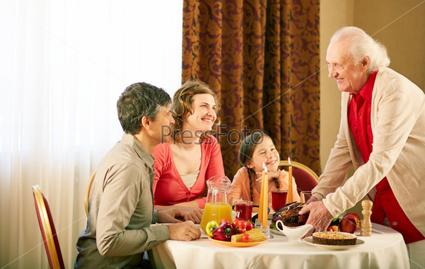 Portrait of happy family sitting at festive table and looking at senior man during Thanksgiving dinner