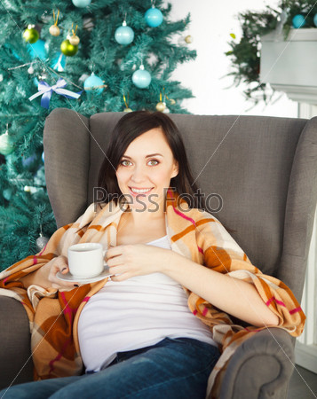 Happy young pregnant woman with cup of tea near the christmas tree siting in armchair