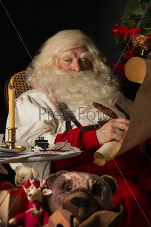 Santa Claus sitting at home and writing on old paper roll to do list with feather pen and ink at night with candle light. Authentic vintage style portrait.