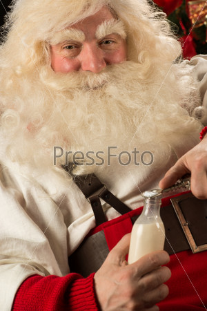 Happy Santa Claus opening glass bottle with milk against Christmas Tree at home.