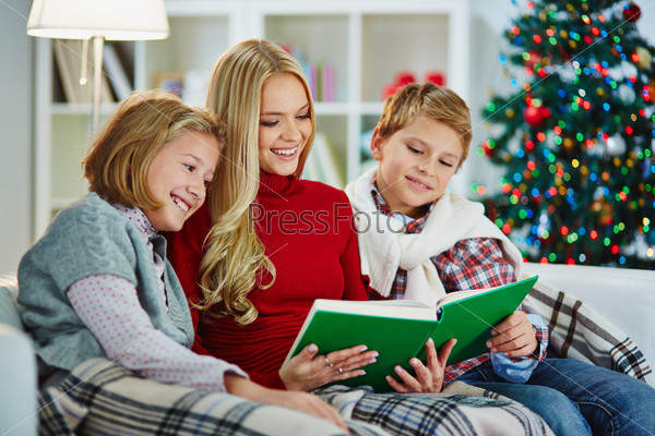 Portrait of pretty woman and her two children reading book on Christmas evening