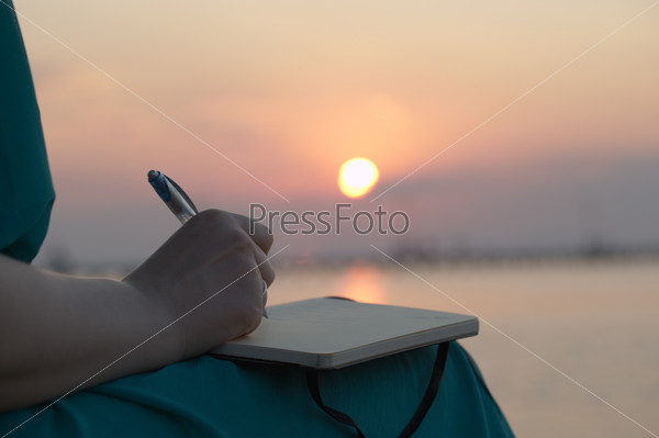 Close Up View Of The Hand Of A Woman Writing In Her Diary At Sunset With The Glowing Orb Of The Sun Reflected Over A Still Ocean