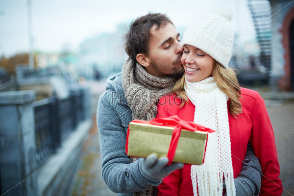 Image of affectionate guy kissing his girlfriend while giving her present outside