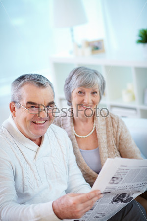Portrait of mature man reading newspaper at home with his wife near by