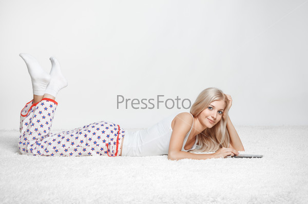 Young blonde woman in pyjamas on white whole-floor carpet reading e-book