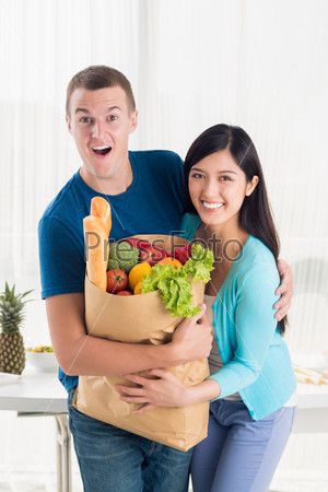 Vertical portrait of an excited couple holding a paper bag with products from local market