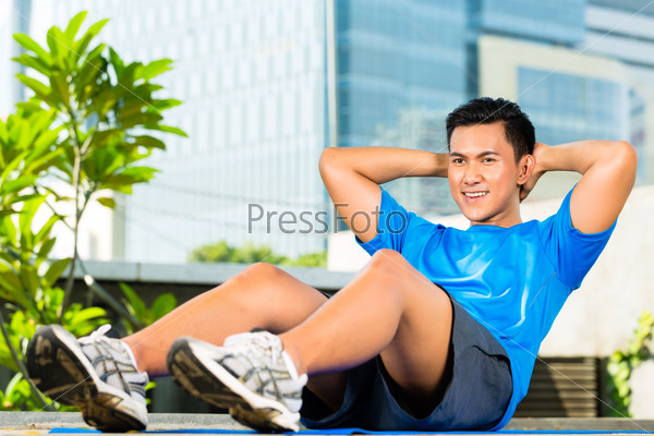 Urban sports - young Asian Indonesian man is doing warming up and sit-ups before running in the city on a beautiful summer day