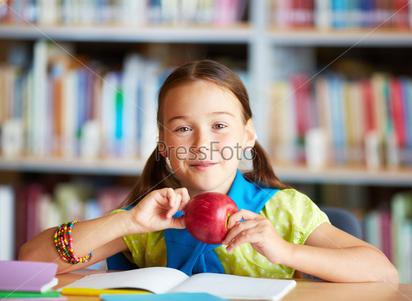 Portrait of happy schoolgirl with apple looking at camera in library
