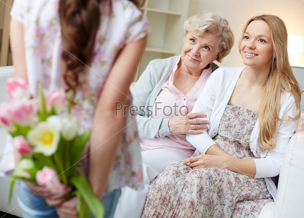 Rear view of little girl holding bunch of flowers behind back with her mother and grandmother looking at her