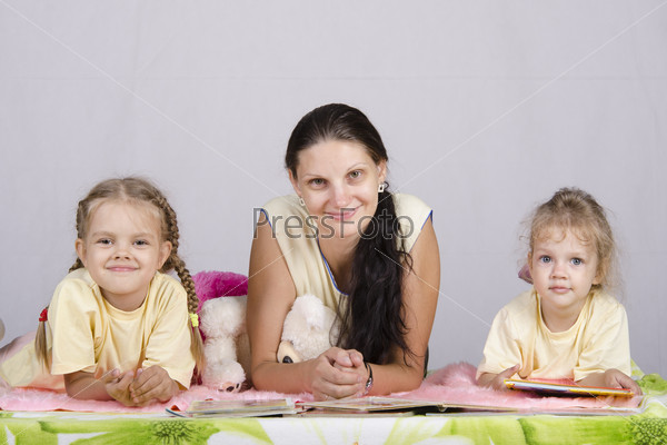 A mother and two daughters lie on the bed and reading the books. They all look to the frame. Studio