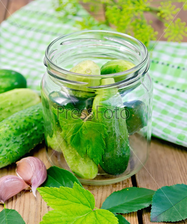 Cucumbers in a glass jar and on the table, garlic, tarragon, dill, napkin, cherries and currants leaves on the background of wooden boards