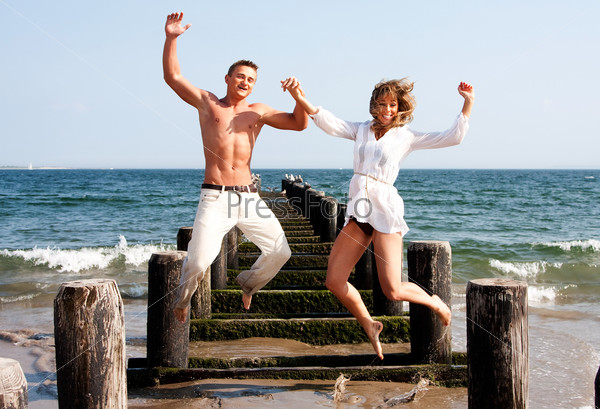 Cute happy couple smiling jumping playfully at the beach with a pier and the ocean water in the background heaving fun