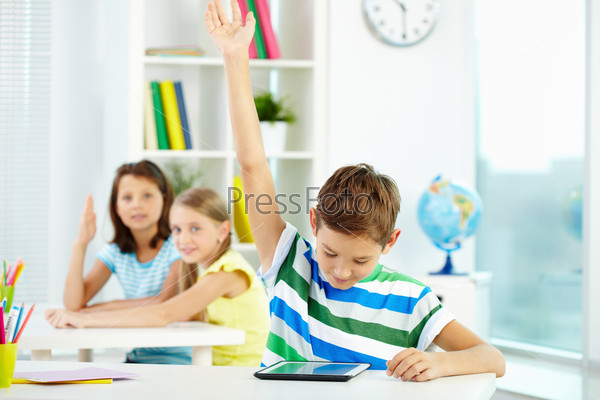 Portrait of smart lad at workplace raising hand and looking at touchpad with his classmates on background