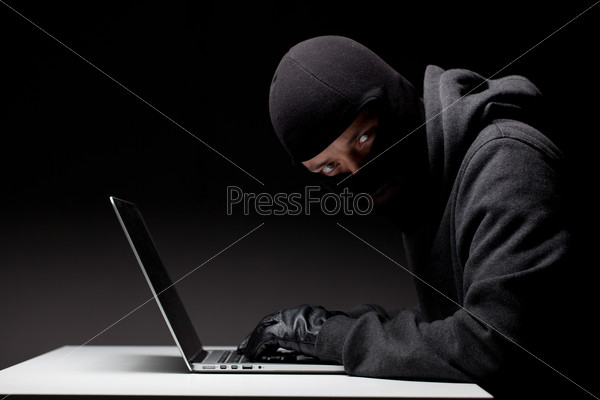 Computer hacker in a balaclava working in the darkness stealing data and personal identity information off a laptop computer