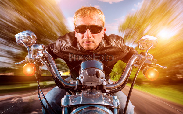 Biker man wearing a leather jacket and sunglasses sitting on his motorcycle looking at the sunset, racing on the road. Filter applied in post-production.
